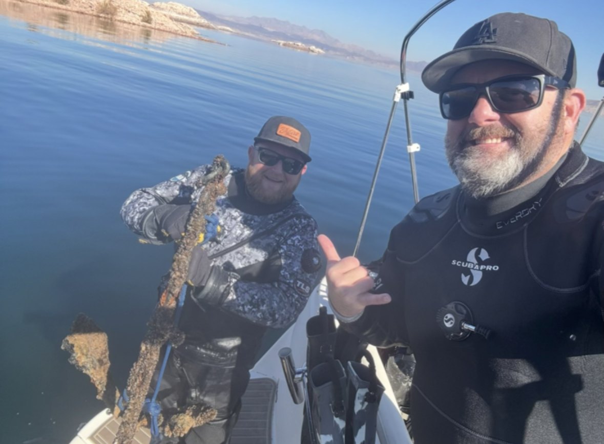 Two divers on a boat, one holding up a recovered anchor chain and the other giving a shaka sign